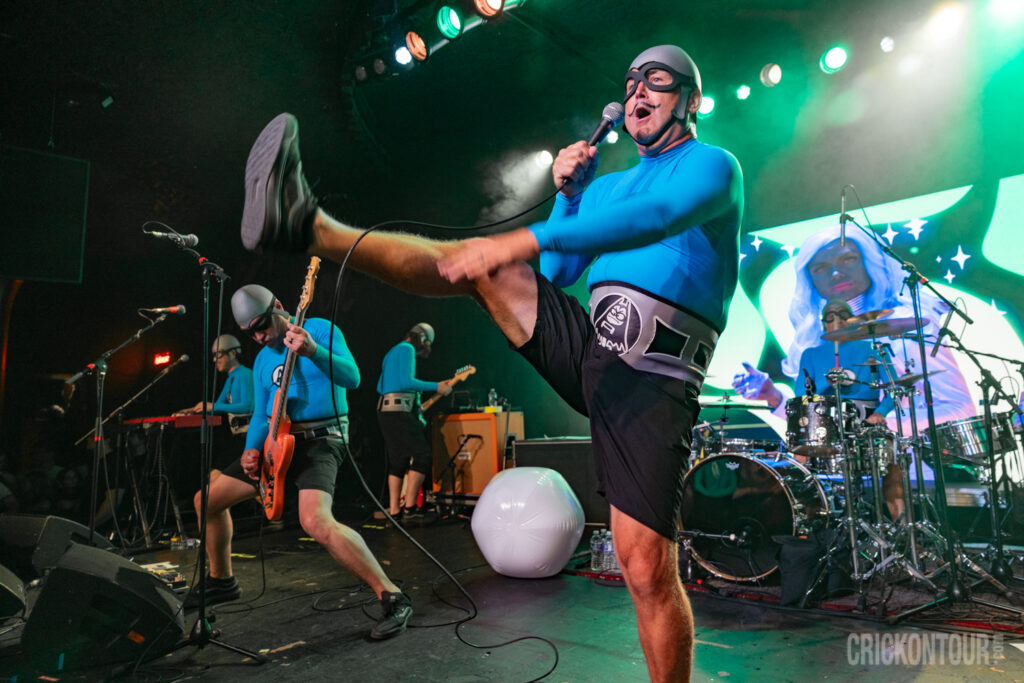 The Aquabats captured mid-performance under green stage lights, with guitarists, drummer, and aM.C. Bat Commander kicking his leg high in the air.