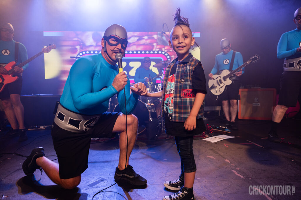A child in plaid vest fist-bumps the masked M.C. Bat Commander while musicians in matching blue outfits play behind them on stage.
