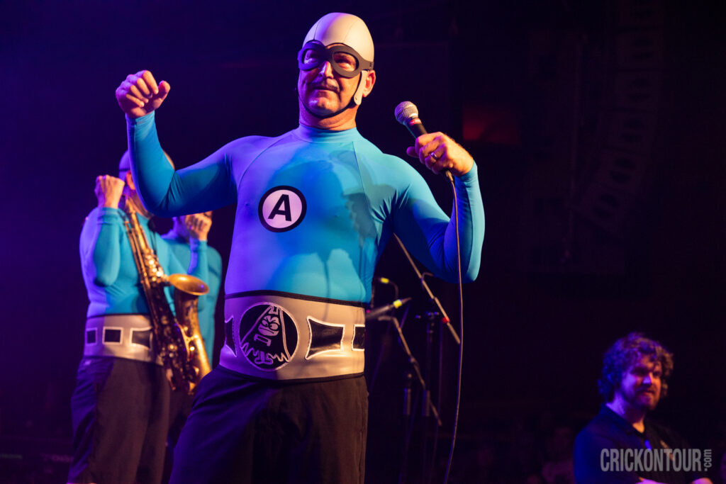 M.C. Bat Commander in aquatic superhero attire performs energetically under purple stage lighting, with a saxophonist and other band members nearby.