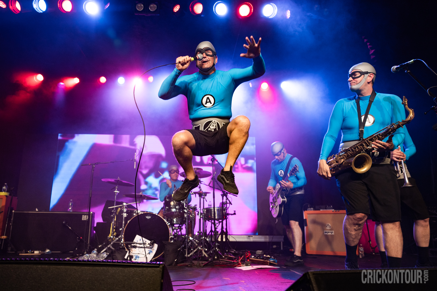 Energetic live concert with colorful red and blue stage lighting, featuring a jumping M.C. Bat Commander of The Aquabats, saxophonist, guitarist, and drummer in costume.