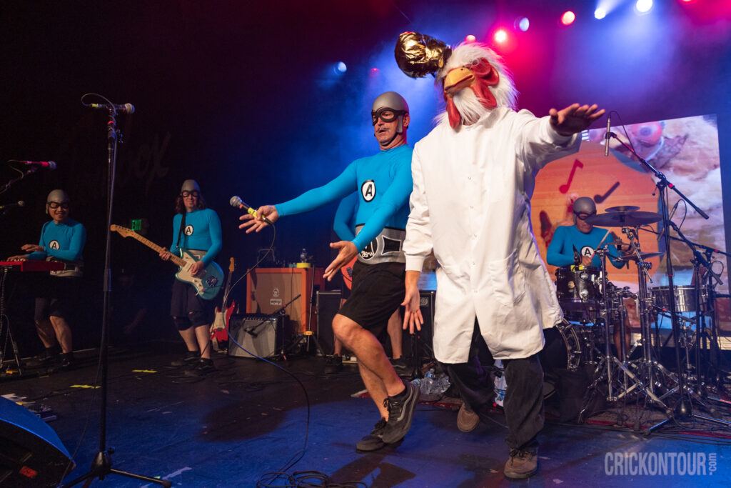 Live rock performance featuring a singer, guitarist, keyboardist, and drummer in matching blue outfits and goggles, plus a costumed chicken figure center stage.