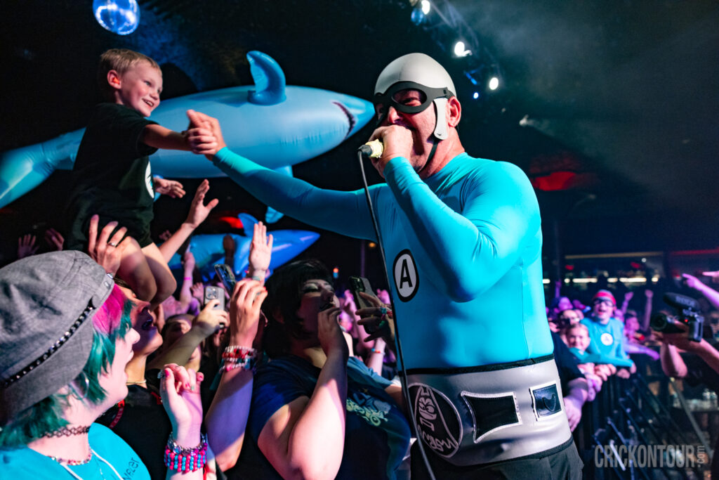 Energetic concert scene with fans filming on phones, inflatable sharks floating overhead, and singer M.C. Bat Commander connecting with a smiling boy in the audience.