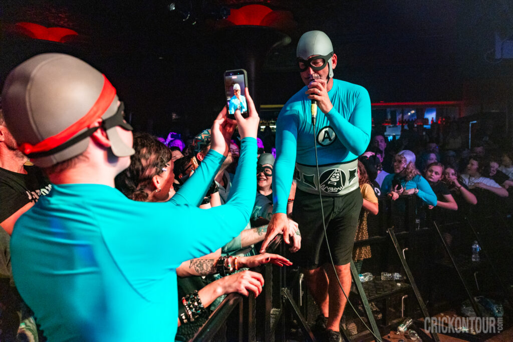 A concert scene where M.C. Bat Commander wearing a a costume emblazoned with an 'A' emblem leans toward an eager crowd of fans in matching caps and goggles.