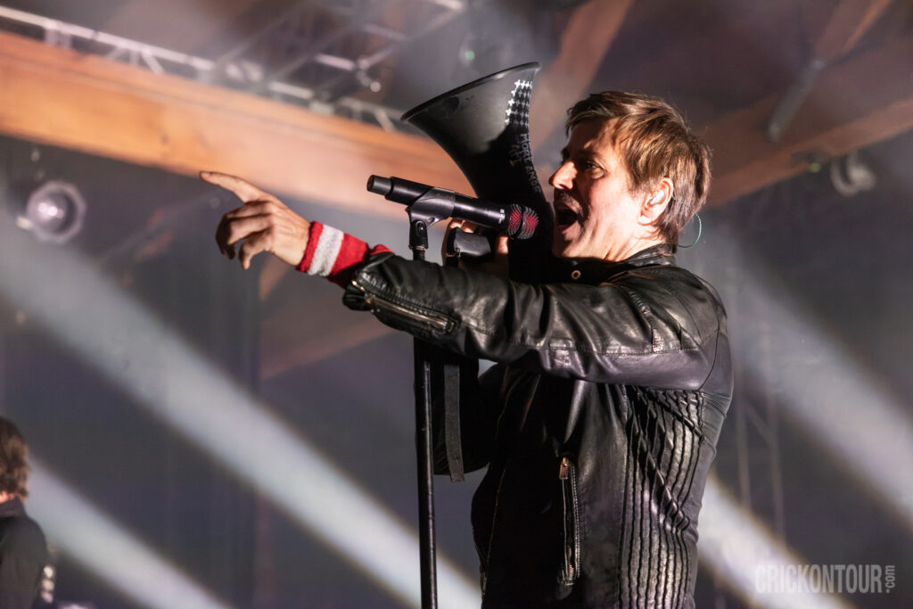 Lead vocalist Raine Maida of Our Lady Peace sings into a megaphone-style mic while pointing outward, wearing a black leather jacket under stage lights.
