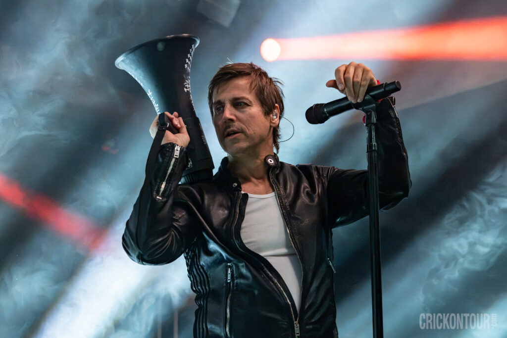 Lead vocalist Raine Maida of Our Lady Peace holds a black megaphone to his shoulder while gripping a mic stand, lit by stage lights and haze.