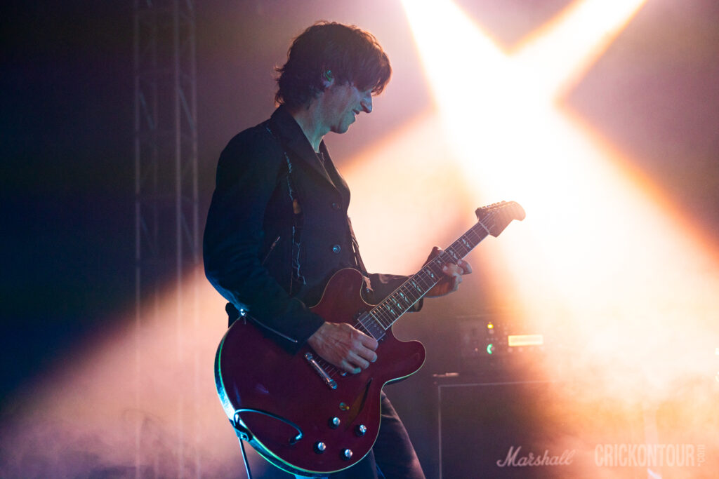 Lead guitarist Steve Mazur of Our Lady Peace plays a red hollow-body guitar on stage, lit by a bright X-shaped stage light.