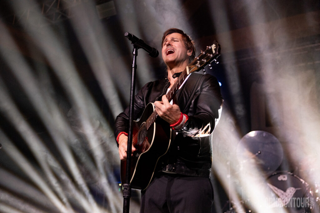 Lead vocalist and acoustic guitarist Raine Maida of Our Lady Peace sings passionately into a mic while playing a black guitar under dramatic stage lighting.