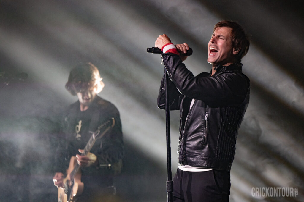 Lead vocalist Raine Maida of Our Lady Peace sings passionately into a mic stand in a black leather jacket, eyes closed, as a guitarist plays behind him in stage haze.