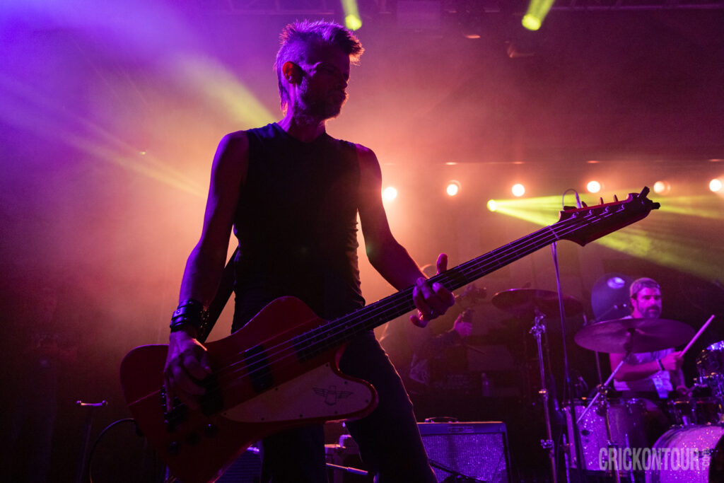 Bass guitarist Brad Vander Ark of The Verve Pipe leans back mid-performance, playing a red bass under purple and yellow stage lights.
