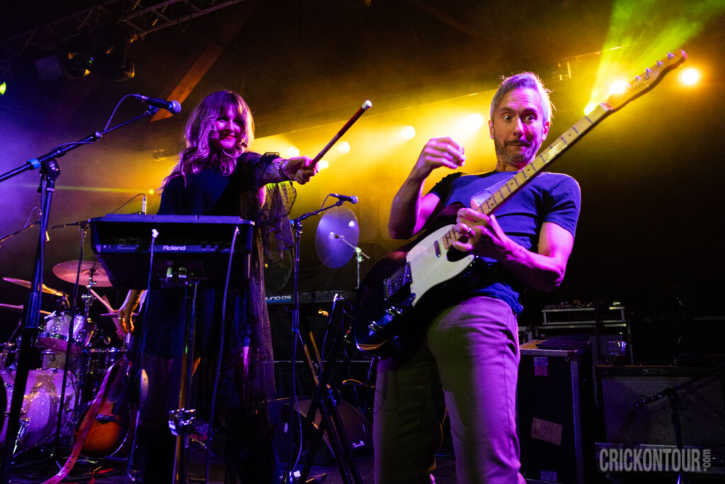 On a colorfully lit concert stage, Daine Hammerle of The Verve Pipe gestures with a drumstick near a Roland keyboard as Brian Vander Ark performs an animated guitar solo in the foreground.