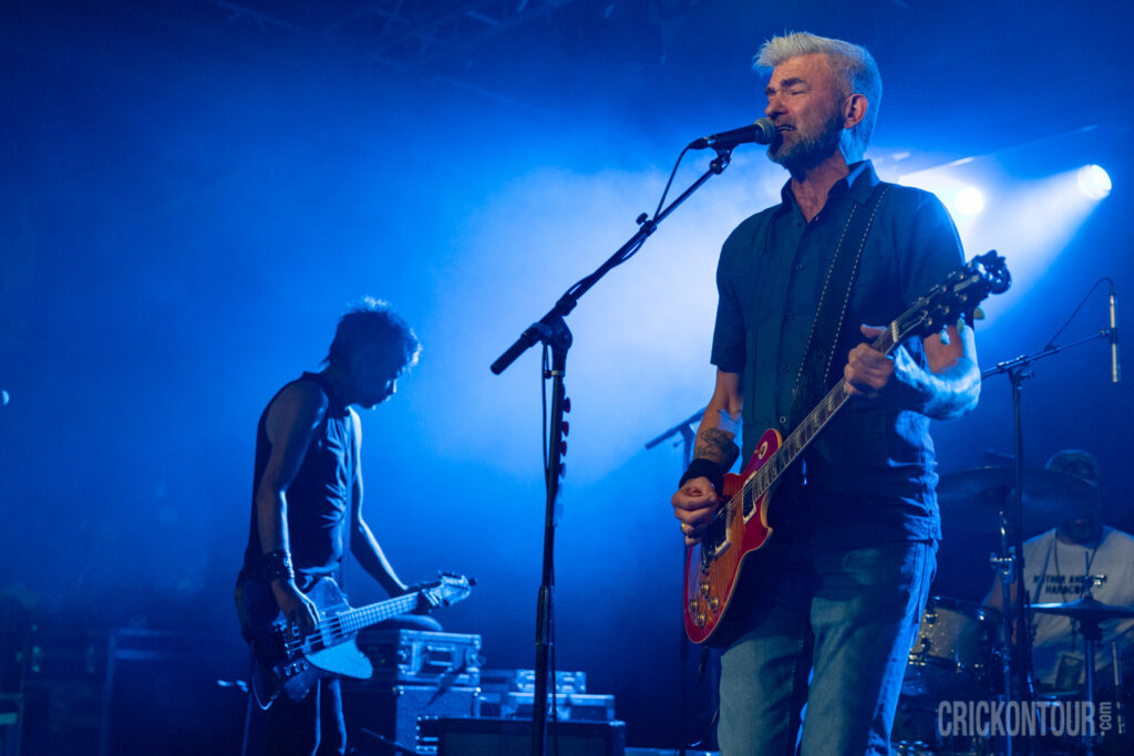 Bassist Brad Vander Ark and drummer Daine Hammerle of The Verve Pipe perform on a smoke-filled, blue-lit stage while a bearded guitarist
Brian Vander Ark sings at the mic in the foreground