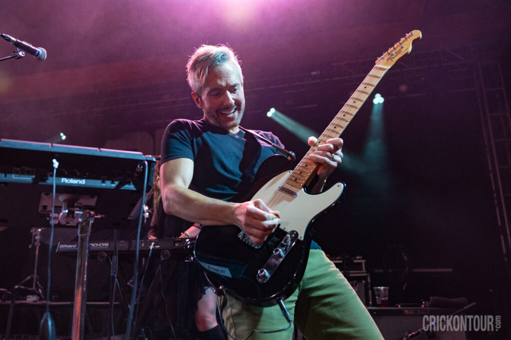 A smiling Lou Musa of The Verve Pipe plays a Fender electric guitar on stage under pink and purple lights, with Roland keyboards behind him.