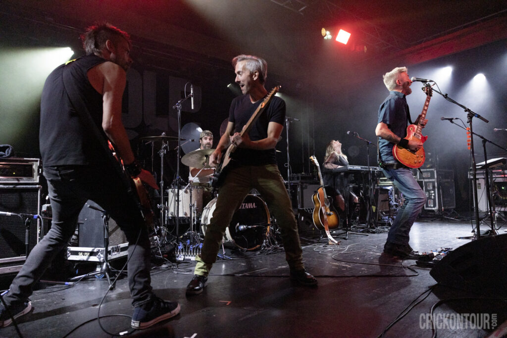 Guitarist
Brian Vander Ark of The Verve Pipe with wide-eyed expression plays a black electric guitar on a stage lit with purple and yellow light, while keyboardist Channing Lee raises a drumstick behind him.