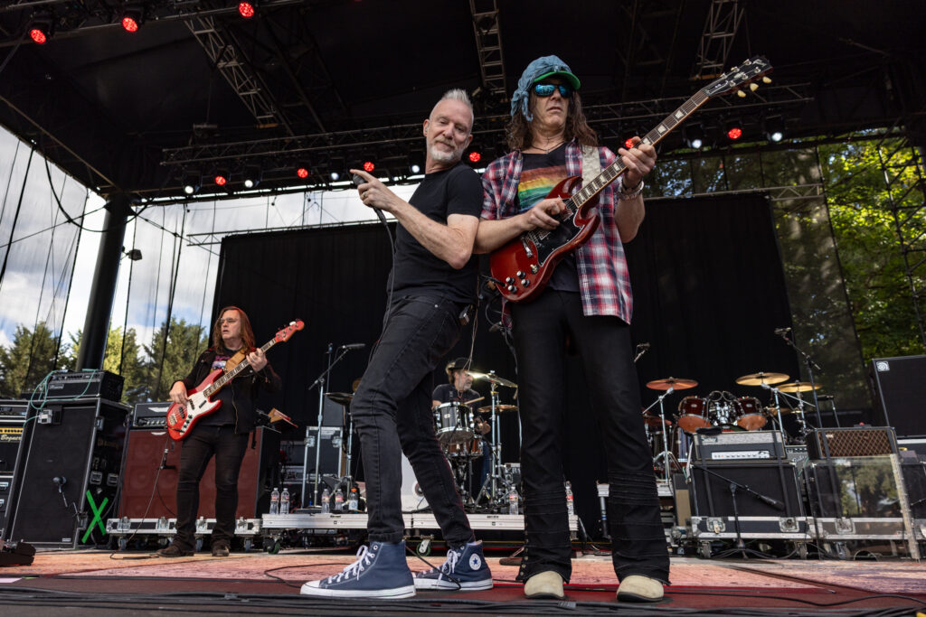 Lead singer Chris Barron and guitarist Eric Schenkman of the Spin Doctors perform on an outdoor stage; Chris gestures toward the camera while Erik plays a red Gibson SG guitar wearing sunglasses and a bandana.