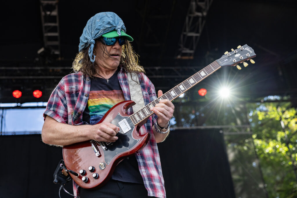 Eric Schenkman, guitarist in the Spin Doctors in a blue bandana, sunglasses, and plaid shirt plays a red Gibson SG electric guitar on an outdoor stage at Marymoor Park in Redmond, Wa.