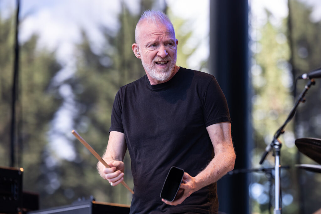 Chris Barron, lead singer of the Spin Doctors in a black t-shirt performs on stage, holding a drumstick and a percussion block.