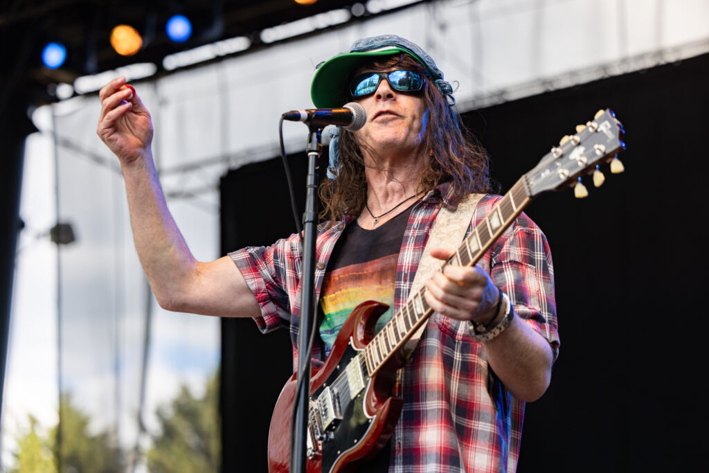 Guitarist Eric Schenkman of the Spin Doctors n a red plaid shirt and rainbow tee performs live, one hand raised with a guitar pick, singing at a mic.