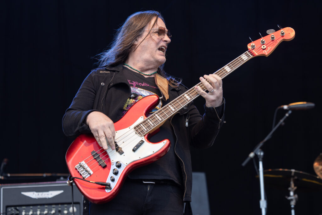 Bassist Jack Daley of the Spin Doctors performs intensely at an outdoor concert while a smiling bandmate stands laughing in the background under stage lighting.