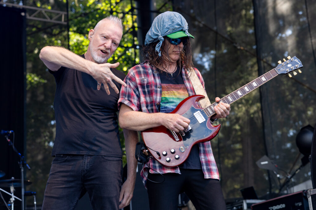 Lead singer Chris Barron and guitarist Eric Schenkman of the Spin Doctors perform on an outdoor stage as both bassist Jack Daley and drummer Aaron Comess jam in the background.