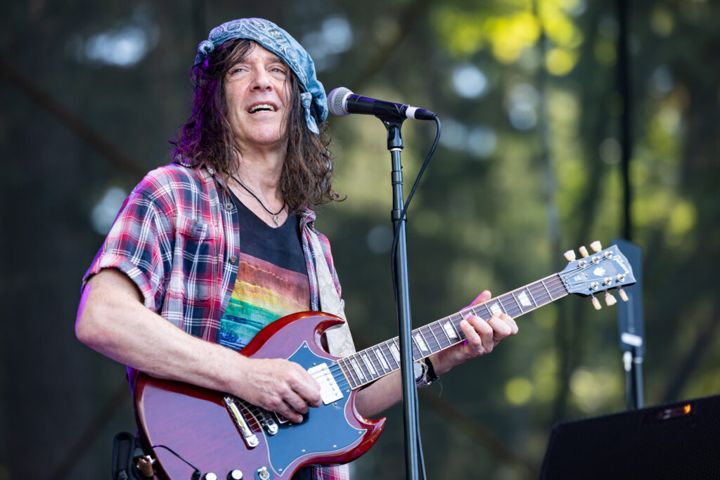 Eric Schenkman of the Spin Doctors performs in a blue bandana, sunglasses, and plaid shirt plays a red Gibson SG electric guitar on an outdoor stage.