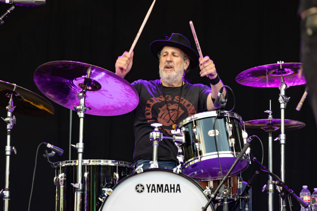 Drummer Aaron Comess of the Spin Doctors performs energetically on drums at a concert, arms lifted with drumsticks, cymbals glowing purple against a dark backdrop.